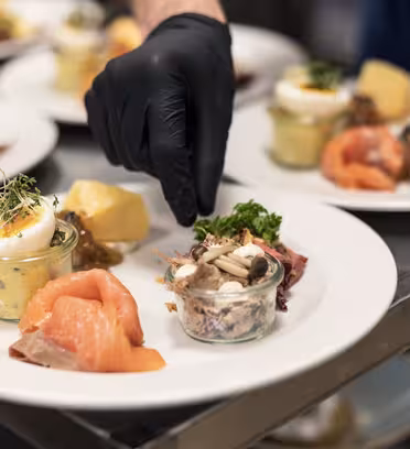 En kok iført sorte handsker pynter tallerkener med forskellige fødevarer, inklusive røget laks, en krukke salat, en skive kartoffel og urter, på en bordplade i rustfrit stål.