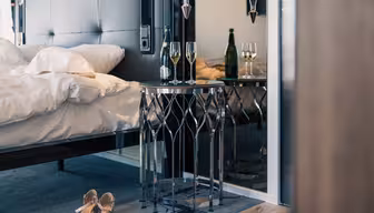 Et luksuriøst hotelværelse har en pænt redt seng, moderne metal sidebord med et geometrisk design, to glas og en flaske champagne. Elegante højhælede sko hviler på gulvet ved siden af sengen.