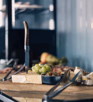 Et bord viser en række forskellige oste på en træplade, med en kniv, der stikker oprejst i én ost. Vindruer og nødder ledsager ostene, og en serveringsbakke af metal er synlig i nærheden. Baggrunden viser flere madvarer lidt ude af fokus.
