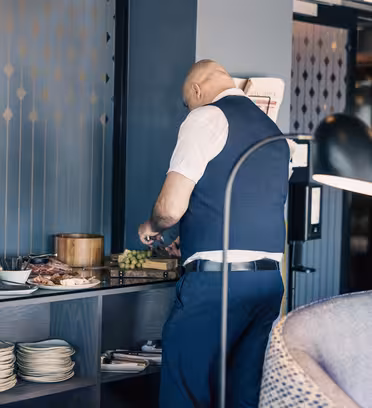 En skaldet mand i hvid skjorte og blå vest er ved at tilberede et fad med mad, inklusive vindruer og kød, på en disk i et moderne værelse. Pladerne er stablet nedenunder. En lampe med en buet hals er i forgrunden.