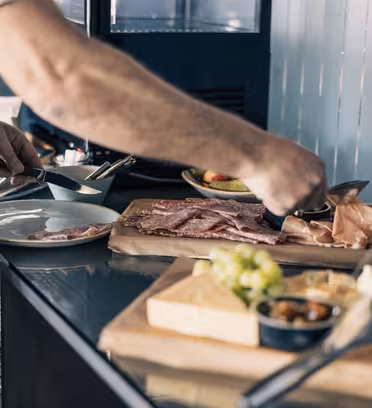 En person i vest og skjorte serverer speget kød på en tallerken ved en buffet. Disken har diverse kød, oste og vindruer på træplader, med andre retter sløret i baggrunden.