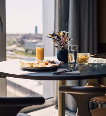 Et rundt bord ved et vindue med udsigt over byen. Den har en tallerken mad, inklusive brød og ost, et glas appelsinjuice, et glas vand, en lille krukke og en vase med blomster. To træstole er placeret rundt om bordet.