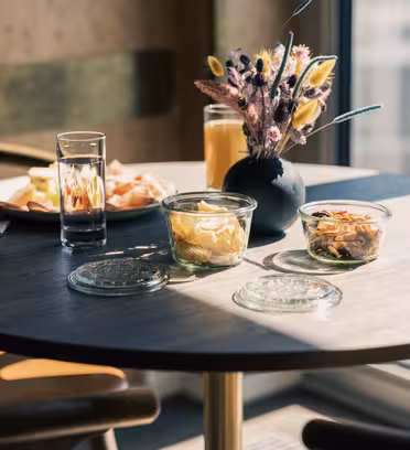 Et solbelyst bord med to overdækkede glasskåle indeholdende mad, et glas vand og et glas appelsinjuice. En lille sort vase rummer et blomsterarrangement. En tallerken med mad er i baggrunden. Indstillingen er ved et stort vindue.