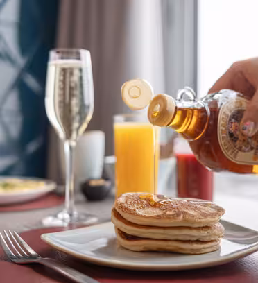 En person hælder sirup over en stak pandekager på en tallerken. Bordet har også et glas champagne, appelsinjuice, en kaffekop og ketchup. En blå geometrisk væg og et vindue med gardiner er i baggrunden.