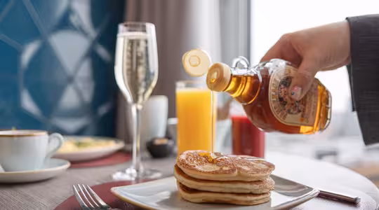 En person hælder sirup over en stak pandekager på en tallerken. Bordet har også et glas champagne, appelsinjuice, en kaffekop og ketchup. En blå geometrisk væg og et vindue med gardiner er i baggrunden.
