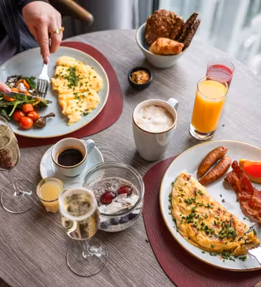 To personer nyder en morgenmad ved et rundt bord. Tallerkenerne byder på omeletter, bacon, pølser og salat. Drikkevarer inkluderer kaffe, appelsinjuice og champagne. Et udvalg af kager ligger i en separat kurv. Indstillingen er godt oplyst af et vindue.