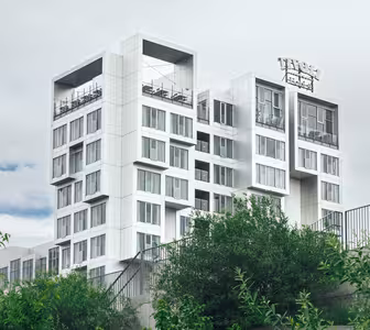 En moderne bygning i flere etager med et unikt geometrisk design med stablede hvide kuber og store vinduer. Det er omgivet af grønt løv og sat mod en overskyet himmel.