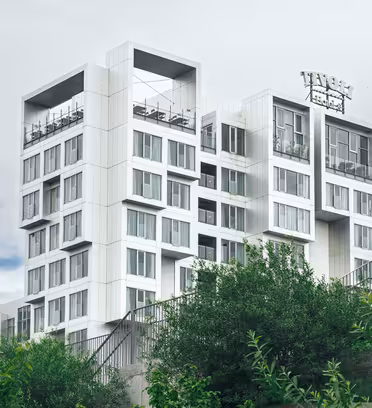 En moderne bygning i flere etager med et unikt geometrisk design med stablede hvide kuber og store vinduer. Det er omgivet af grønt løv og sat mod en overskyet himmel.