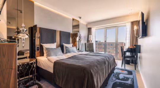 Moderne hotelværelse med stor seng, gråt og hvidt sengetøj og mørkegrå lænestol. Der er elegante sideborde med lamper, et vægmonteret tv, et skrivebord med en stol og store vinduer med udsigt over byen. Indretningen er slank og moderne.