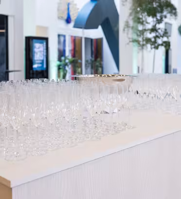 Et stort antal tomme champagneglas pænt arrangeret på et lyst træbord i et lyst, moderne rum. Der er høje grønne planter og digitale displays synlige i baggrunden.