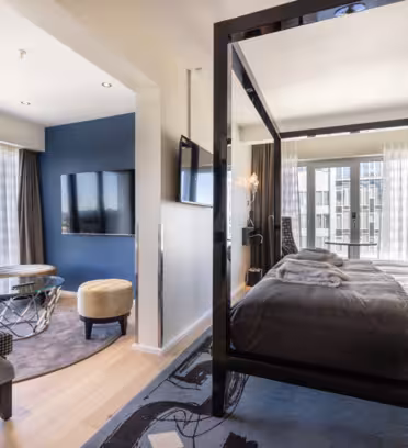 Lys hotelsuite med en moderne himmelseng, gråt sengetøj og matchende puder. Et opholdsområde omfatter et rundt glasbord og behagelige stole. Store vinduer slipper naturligt lys ind, og der er monteret fladskærms-tv på væggen.