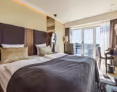 Moderne hotelværelse med to senge dækket af grå tæpper og guldpuder. Værelset har en hovedgærde i træ, gulv-til-loft-vinduer med gennemsigtige gardiner, udsigt over byen, lille skrivebord med stol og en stilfuld lampe. Lyse vægge og trægulve.
