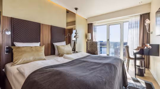 Moderne hotelværelse med to senge dækket af grå tæpper og guldpuder. Værelset har en hovedgærde i træ, gulv-til-loft-vinduer med gennemsigtige gardiner, udsigt over byen, lille skrivebord med stol og en stilfuld lampe. Lyse vægge og trægulve.