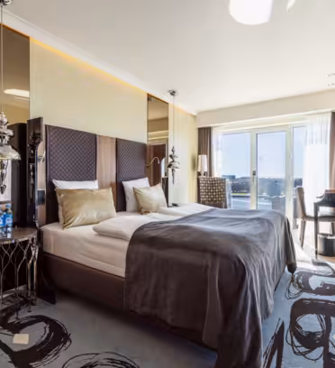 Moderne hotelværelse med dobbeltseng, gråt tæppe og to guldpuder. Værelset har spejlvægge, et dekorativt sidebord, to lamper og et stort vindue med udsigt. Et skrivebord med en stol er ved vinduet, oplyst af naturligt lys.