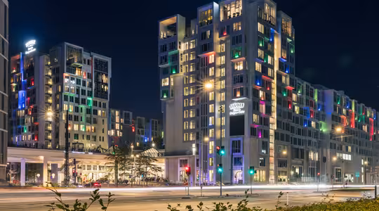 Natscene af et moderne hotelkompleks med flere bygninger. Strukturerne er stærkt oplyste og byder på farverigt oplyste vinduer. En vej med kørende billygter er i forgrunden, og gadelygter langs vejen.