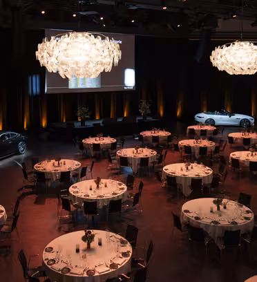 En svagt oplyst festsal med runde borde til spisning. Elegante lysekroner hænger fra loftet. To biler vises på scenen, og en stor skærm er synlig i baggrunden. Atmosfæren er formel og sofistikeret.