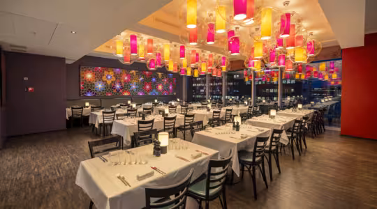Et moderne restaurantinteriør med pænt arrangerede borde dækket af hvide duge og dækket med tallerkener og glas. Loftet er prydet med farverige, oplyste lanterner. En stor, dekorativ væg har livlige mønstre.