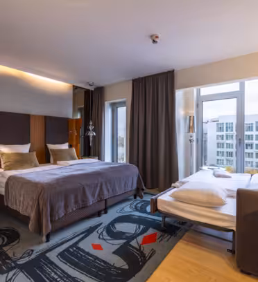 Et moderne hotelværelse med to senge; den ene queen-size med gråt og beige sengetøj, den anden enkelt med hvidt sengetøj. Rummet har store vinduer, grå gardiner, et farverigt tæppe og et skrivebord med en stol. Et bybillede er synligt udenfor.