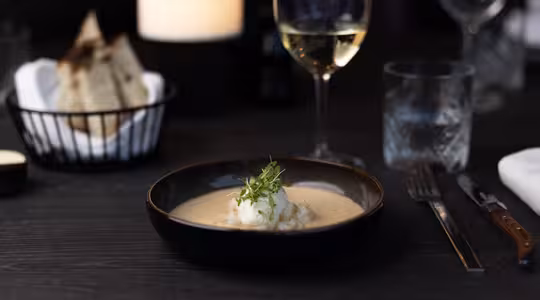 En skål cremet suppe pyntet med krydderurter stilles på et mørkt bord. Et glas hvidvin stilles ved siden af. I baggrunden tilføjer en kurv med brød og et stearinlys den elegante spisestue.