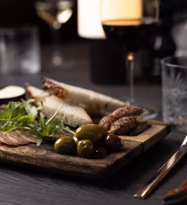På et mørkt bord står en træplade med skiveskåret brød, charcuteri, oliven og rucola. Et glas rødvin og et glas vand med is er i nærheden, sammen med en kniv og gaffel pakket ind i en serviet. En sløret baggrund har flere vinglas.
