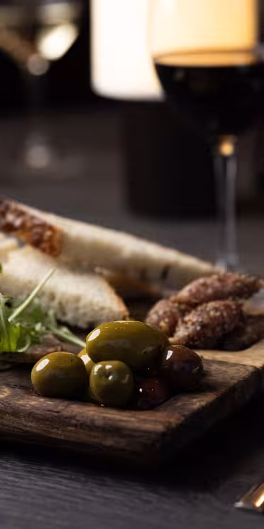 På et mørkt bord står en træplade med skiveskåret brød, charcuteri, oliven og rucola. Et glas rødvin og et glas vand med is er i nærheden, sammen med en kniv og gaffel pakket ind i en serviet. En sløret baggrund har flere vinglas.