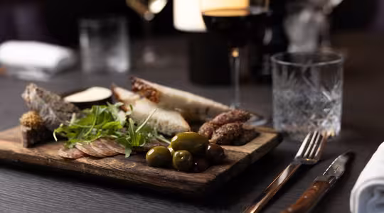 På et mørkt bord står en træplade med skiveskåret brød, charcuteri, oliven og rucola. Et glas rødvin og et glas vand med is er i nærheden, sammen med en kniv og gaffel pakket ind i en serviet. En sløret baggrund har flere vinglas.