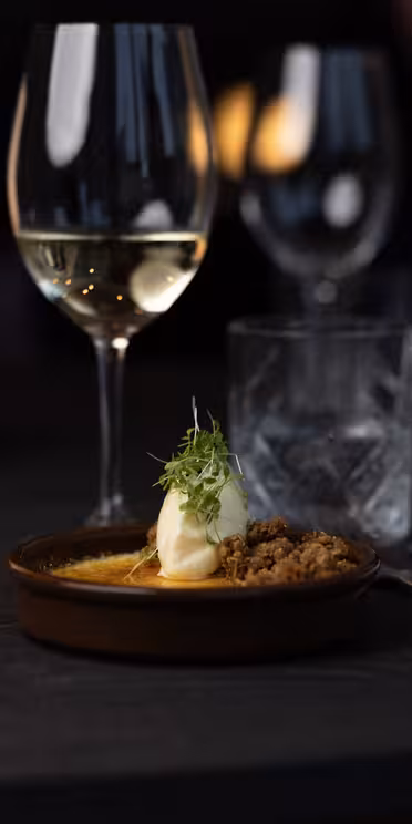 En gourmetret med wienerbrød og flødepynt står på et mørkt træbord, ledsaget af et glas hvidvin, en isdrik, redskaber og en rullet serviet. En lille lampe oplyser blidt omgivelserne og skaber en elegant spisestemning.