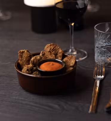En skål sprøde svampe ved siden af et lille fad rød dipsauce på et mørkt træbord. I nærheden er et fyldt vinglas, et tomt vandglas, en gaffel og en kniv. Atmosfæren er svag med blød belysning.
