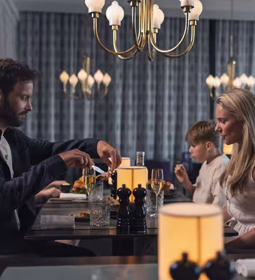En familie på tre spiser i en restaurant med elegante lysekroner, der oplyser rummet. Manden og kvinden er i snak, mens drengen nyder sit måltid. Bordet er dækket med glas og peberkværne.
