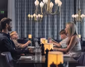 En familie på fire sidder ved et restaurantmiddagsbord. Forældrene er over for hinanden, med to børn siddende mellem dem. De er engageret i samtale og omgivet af elegante lysekroner og varm belysning.