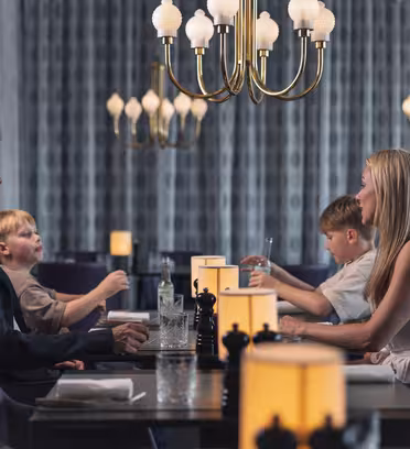 En familie på fire sidder ved et restaurantmiddagsbord. Forældrene er over for hinanden, med to børn siddende mellem dem. De er engageret i samtale og omgivet af elegante lysekroner og varm belysning.