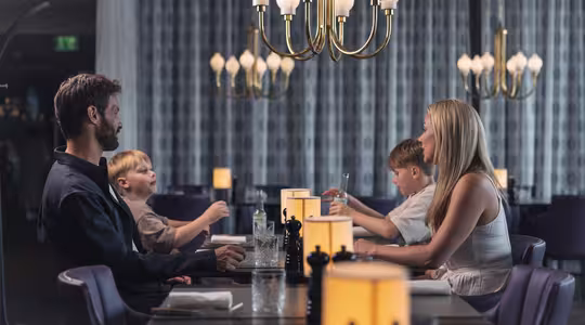 En familie på fire sidder ved et restaurantmiddagsbord. Forældrene er over for hinanden, med to børn siddende mellem dem. De er engageret i samtale og omgivet af elegante lysekroner og varm belysning.