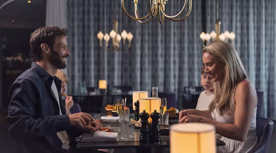 Et par sidder overfor hinanden ved et restaurantbord og smiler og sludrer. Manden sidder til venstre, kvinden til højre. Rummet er blødt oplyst med elegante lysekroner og et mønstret gardin i baggrunden.
