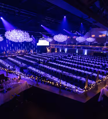 En stor eventhal er sat til en formel sammenkomst. Lange selskabsborde er prydet med borddækning og omgivet af stole. Blå og lilla belysning skaber en elegant atmosfære, med dekorative lysekroner hængende fra loftet.