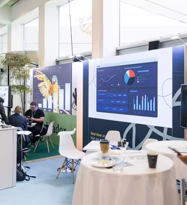 En moderne udstillingshal med små boder, informationsudstillinger og mennesker, der er engageret i samtale. En digital skærm viser datadiagrammer og grafer. En person i forgrunden holder en mikrofon, mens andre sidder og interagerer ved forskellige kabiner.
