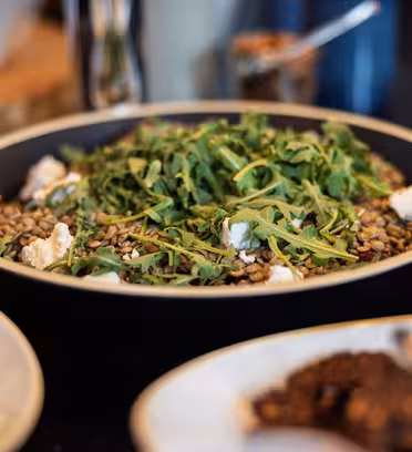 En salat i en sort skål med linser, rucola og stykker ost. Fadet står på et bord med andre tallerkener og skåle, sløret i baggrunden, hvilket tyder på en spise- eller buffetindstilling.