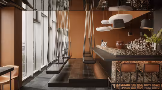 En moderne bar med gyngesæder langs disken. Baren har en stenlignende finish og varm-tonet belysning med store vinduer på den ene side. Hængende hvide og sorte pendellamper tilføjer kontrast til det moderne design.