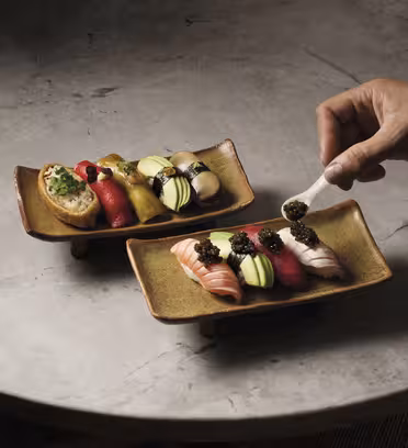 En hånd placerer forsigtigt kaviar på et stykke sushi på et rektangulært keramisk fad. Begge skåle består af forskellige farverige sushistykker arrangeret på en struktureret stenoverflade.
