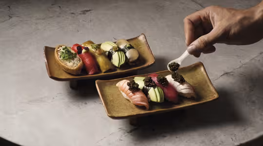 En hånd placerer forsigtigt kaviar på et stykke sushi på et rektangulært keramisk fad. Begge skåle består af forskellige farverige sushistykker arrangeret på en struktureret stenoverflade.