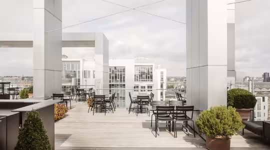 En moderne tagterrasse har adskillige sorte borde og stole på et træterrasse. Potteplanter er spredt ud over det hele, og baggrunden afslører en udsigt over byens bygninger under en overskyet himmel.
