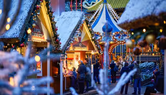 Festligt udendørs julemarked med snedækkede træboder pyntet med lys og guirlander. Folk samles omkring en kaffestand. Dekorative lanterner og en karrusel er synlige i baggrunden.