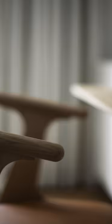 En træstol med buet ryglæn er placeret ved siden af et træbord. Bordkanten og en vase med grønne blade er synlige. Baggrunden er blødt sløret, hvilket tyder på naturligt lys, der kommer gennem et vindue.