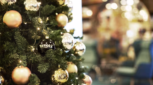 Et pyntet juletræ prydet med guld og pink ornamenter, inklusive en sort ornament mærket "Tivoli." Baggrunden er blødt sløret og viser antydninger af en festlig indendørs indstilling med blød belysning.