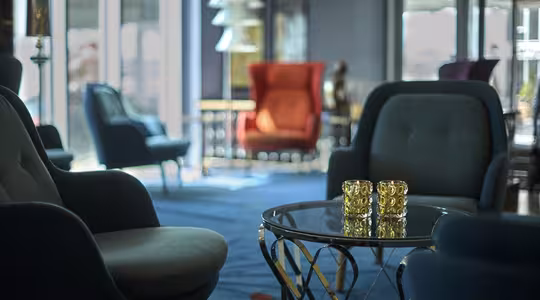 Et loungeområde med moderne indretning med flere lænestole og et glas sofabord med to gulmønstrede glas. Naturligt lys filtrerer ind gennem store vinduer, og en rød lænestol ses i baggrunden. Tæppet er dybt blåt.