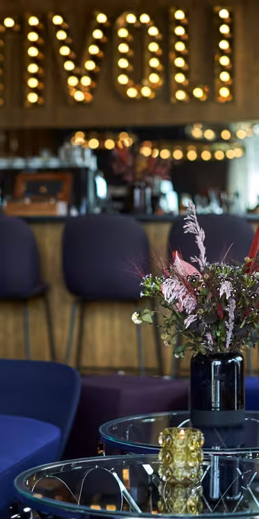 Et hyggeligt loungeområde med farverige stole og et stilfuldt blomsterarrangement på et glasbord. Bagved har en bar en trævæg med oplyste bogstaver, der staver "TIVOLI", hvilket skaber en varm og indbydende atmosfære.