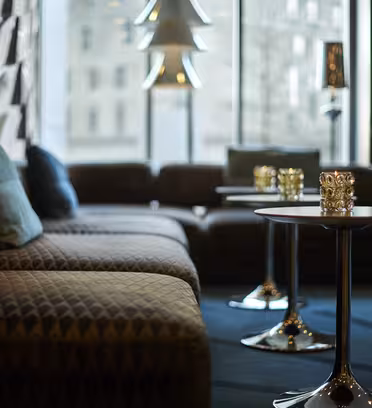 Luksuriøst loungeområde med mønstrede sofaer og små runde borde toppet med dekorative lys. Store vinduer giver naturligt lys og viser et sløret bybillede udenfor. En unik lampe og moderne indretning fuldender de stilfulde rammer.