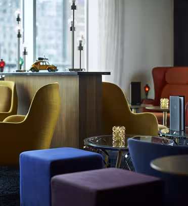En moderne lounge med farverige møbler med sennepsgule og røde stole, blå og lilla ottomaner og runde glasborde. Dekorative genstande omfatter en lille legetøjsbil og tændte stearinlys. Store vinduer giver udsigt til byens bygninger udenfor.