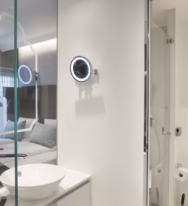 Moderne hotelværelse med et badeværelse med glasvægge med et cirkulært spejl og håndvask. Hovedområdet omfatter en seng, natbord og lampe, med lys, der strømmer gennem store vinduer dækket af rene gardiner.