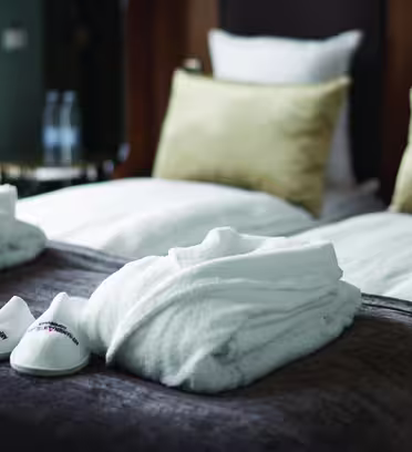 Et hyggeligt hotelværelse med en pænt redt seng med to puder og et tekstureret tæppe. To bløde hvide badekåber og et par hvide hjemmesko er placeret på sengen, hvilket tilføjer et strejf af luksus.