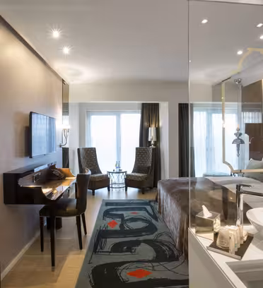Et moderne hotelværelse med dobbeltseng, skrivebord med stol og væghængt tv. To polstrede stole vender ud mod et vindue med gennemsigtige gardiner. Værelset har stilfuld indretning, en spejlvæg og et synligt håndvaskområde med toiletartikler.